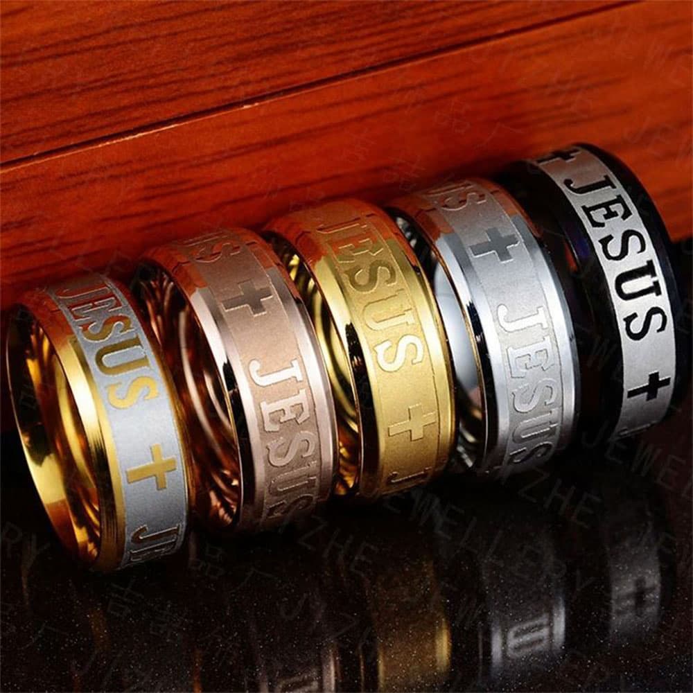 Close-up reflection view of 8mm stainless steel Jesus cross bands resting on a dark reflective surface against a wooden backdrop, highlighting the detailed religious engraving.
