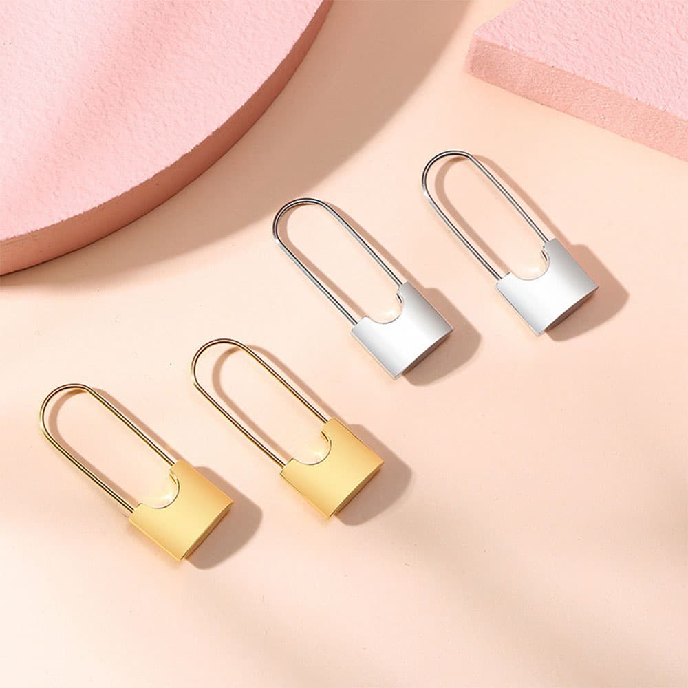 Flat lay view of gold and silver padlock dangle earrings on a beige and pink geometric background, showing a polished reflective finish.