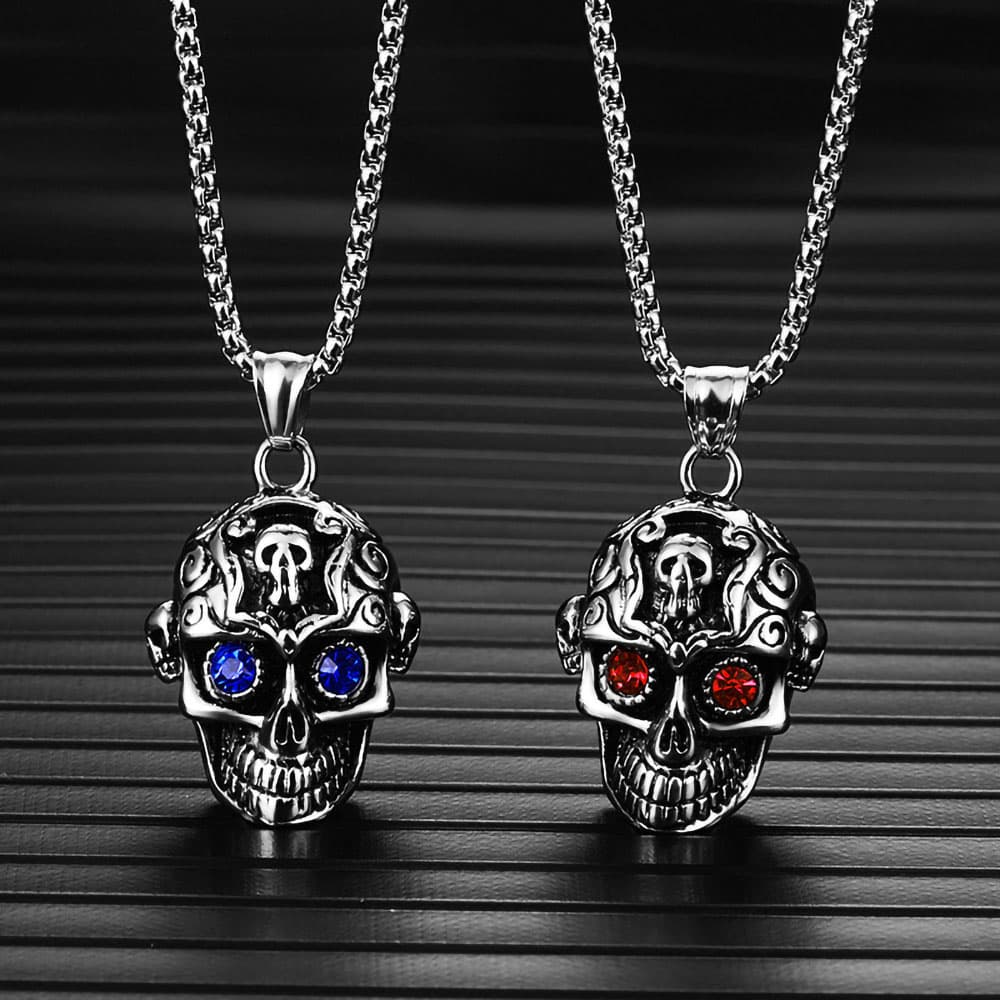 Front view of two men's stainless steel carved skull pendants with blue and red crystal eyes on a dark background.